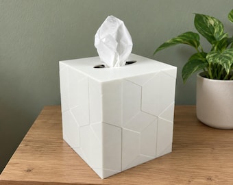 May include: A white square tissue box cover featuring a geometric pattern of raised lines on its exterior surfaces. A single white tissue is pulled through the oval-shaped opening at the top of the box, extending upward from the center.