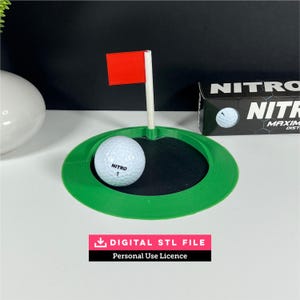 Peut inclure: Une coupe de golf en plastique vert avec un centre noir et un mât blanc surmonté d'un drapeau rouge. Une balle de golf blanche Nitro 1 est placée dans la coupe. À côté se trouve une boîte Nitro noire. Texte : DIGITAL STL FILE Personal Use Licence.