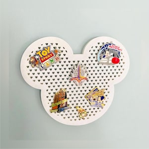 May include: White Mickey Mouse-shaped pin display board with heart-shaped cutouts. The board holds several colourful enamel pins, including designs from Toy Story Land, Space Mountain, and Tower of Terror. The pins are attached to the board.
