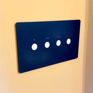 May include: A navy blue rectangular control panel with four white circular buttons. The panel has small white icons above each button. The panel is mounted on a light yellow wall.