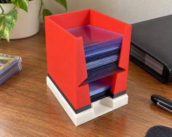 Puede incluir: Una caja de almacenamiento de tarjetas de plástico rojo y blanco con base rectangular y parte superior abierta. La caja presenta una franja horizontal negra que separa la sección superior roja de la inferior blanca para fundas de tarjetas.