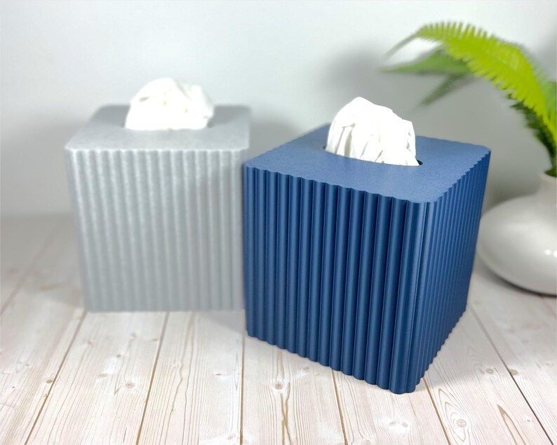 May include: Two square tissue box covers with a vertical ribbed texture. One cover is light gray and the other is dark blue. Each box has a rectangular opening at the top with white tissues protruding from the center of the containers.