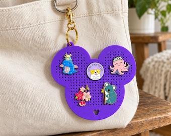 May include: A purple, mouse-shaped silicone pin display board with a gold-tone metal clasp attached to a beige canvas tote bag. The board features five enamel pins: a blue dragon, a round Stay Pawsitive dog pin, a pink octopus, cherry blossoms, and a koala.
