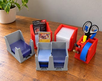 Puede incluir: Un conjunto de organizadores de plástico rojo y gris para tarjetas coleccionables y material de oficina. Incluye bandejas para fundas, top loaders y bolsas, además de un organizador de escritorio rojo con tijeras, cinta y bolígrafos.