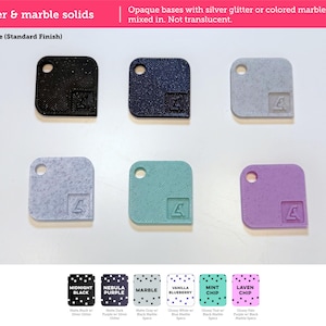 May include: Six square tiles with rounded corners and a circular hole in one corner. Each tile features a small embossed logo. The colors include black, dark purple, gray, light blue, teal, and light purple, with glitter or marble speckles.