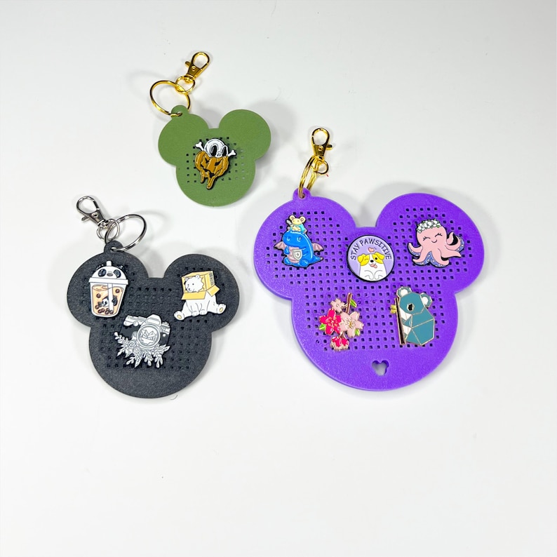 May include: Three mouse-shaped pegboard keychains in olive green, black, and purple. The black board holds three pins: a panda boba tea, a bear in a box, and a camera. The purple board holds four pins: a blue dragon, a dog with text STAY PAWSITIVE, an octopus, and flowers.