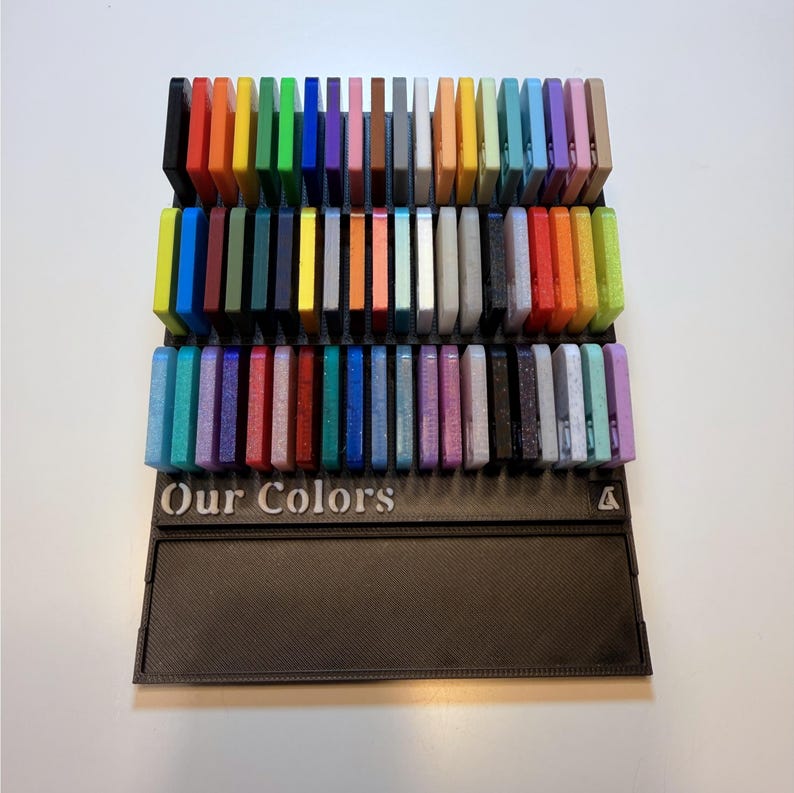 May include: A black plastic display stand featuring three horizontal rows of rectangular color swatches in various shades, including red, blue, green, yellow, and purple. The base of the stand has the text Our Colors embossed in white lettering.
