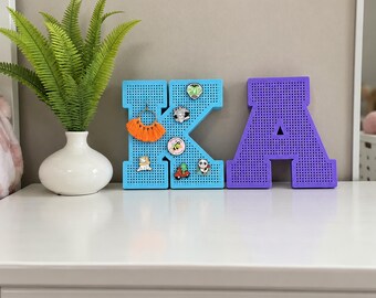 May include: A light blue letter K and a purple letter A, both featuring a perforated pegboard surface. The blue letter K is decorated with several small enamel pins and an orange tassel earring. A green fern plant sits in a white vase.