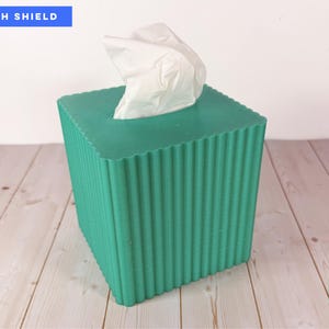 May include: A square emerald green tissue box cover with a vertical ribbed texture and a scalloped top edge. A white tissue protrudes from the center opening. The text WITH SHIELD appears in the top left, and EMERALD GREEN is in the bottom right.