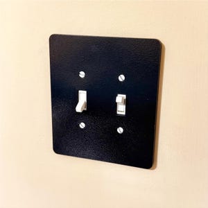 Könnte beinhalten: Eine schwarze, strukturierte Lichtschalterplatte mit zwei weißen Lichtschaltern und vier silbernen Schrauben. Die Lichtschalter befinden sich in der Ein- und Aus-Position. Die Platte ist an einer beigen Wand montiert.