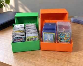 Puede incluir: Una caja de almacenamiento de plástico verde brillante llena de tarjetas coleccionables y una etiqueta blanca que dice Reverse Holos, junto a una caja naranja con dos filas de tarjetas en fundas protectoras transparentes.