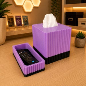 Può includere: Un copriscatola per fazzoletti quadrato di colore viola con una trama a coste e una base nera, abbinato a un vassoio rettangolare coordinato viola e nero contenente un telecomando nero. La scatola contiene dei fazzoletti bianchi.