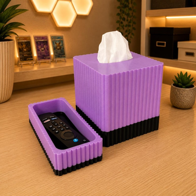 May include: A square purple tissue box cover with vertical ribbed detailing and a black base, featuring a white tissue protruding from the top. Beside it sits a matching rectangular purple tray with a black base containing a black remote control.