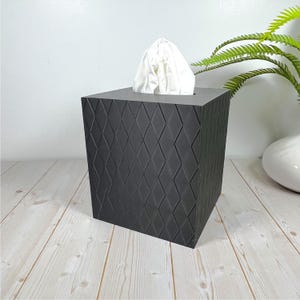 May include: A square, black tissue box with a diamond pattern. White tissues are visible at the top. The box is made of a solid material and sits on a light wood surface. A fern and a white vase are in the background.