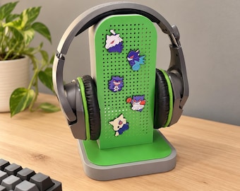 May include: A gray and green headphone stand featuring a perforated green panel decorated with five small character charms. The stand holds a pair of gray over-ear headphones with black ear cushions and green inner accents.