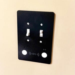 May include: A black rectangular light switch plate with two toggle switches and two round white buttons. The plate has the text "((NFC))" printed on it. The switches are in the on and off positions.