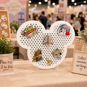 Può includere: Un espositore per spille bianco a forma di testa di Topolino con una griglia a cuori, che ospita cinque spille smaltate colorate, inclusi i design di Toy Story Land, Space Mountain e Tower of Terror, su due supporti in acrilico trasparente.