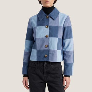 Könnte beinhalten: Eine kurz geschnittene Jacke mit einem Patchwork-Design aus verschiedenen blauen Wildlederquadraten. Das Kleidungsstück verfügt über einen klassischen Kragen, lange Ärmel und eine Knopfleiste mit fünf dunkelbraunen Knöpfen auf der Vorderseite.