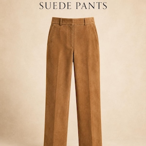 May include: Tan suede trousers featuring a high-waisted design with a front zip closure, belt loops, and side pockets. The text "DESIGN YOUR OWN SUEDE PANTS" is printed in black above the garment, which has a straight-leg cut and a smooth texture.