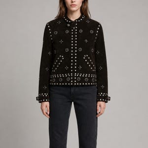 May include: A dark brown suede jacket featuring a round neckline, a front zip fastening, and silver-toned metal stud embellishments arranged in circular and linear patterns across the front, sleeves, and hem, paired with dark grey denim jeans.