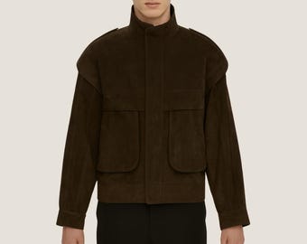 May include: A dark brown suede jacket with a high collar and two large front pockets. The jacket has a boxy silhouette and a slightly padded shoulder design. The sleeves are long, and the jacket is worn over black pants.