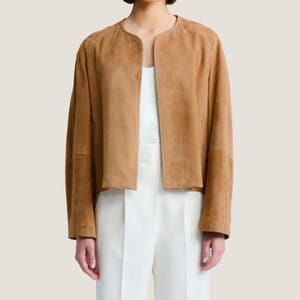 Puede incluir: Chaqueta de ante marr&oacute;n claro con cuello redondo y mangas largas. La chaqueta est&aacute; abierta por delante, revelando una parte superior y un pantal&oacute;n blancos. La chaqueta tiene una longitud recortada y un aspecto suave y texturizado. El estilo general es minimalista y elegante.