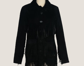Puede incluir: Chaqueta negra de ante con mangas largas y cuello clásico. La chaqueta presenta flecos decorativos en el pecho, los bolsillos y el dobladillo. Tiene cierre de botones y se exhibe en un maniquí.