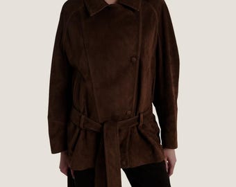 Op de afbeelding: Een bruine suède jas met een dubbelborstig ontwerp en een bijpassende riem. De jas heeft een reverskraag en lange mouwen. De riem sluit aan de taille, waardoor een flatterend silhouet ontstaat. De jas heeft een rijke, warme bruine kleur.