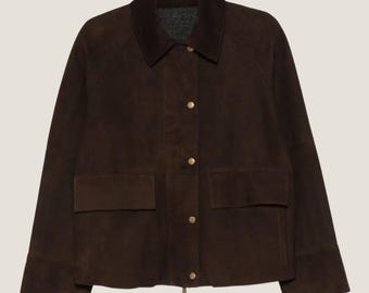 Könnte beinhalten: Dunkelbraune Wildlederjacke mit Kragen und zwei Vordertaschen. Die Jacke hat goldfarbene Knöpfe und einen Reißverschluss. Der Kragen ist dunkler braun und die Jacke hat lange Ärmel.