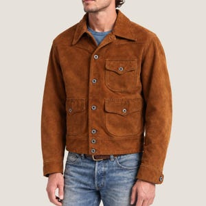 May include: A tobacco brown suede jacket featuring a pointed collar, a button-front closure, and four front patch pockets with buttoned flaps. The jacket has long sleeves with buttoned cuffs and is worn over a blue shirt and blue denim jeans.