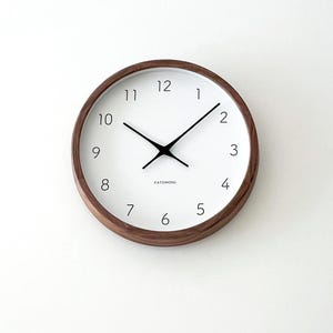 May include: A round wall clock with a dark brown wooden frame and a white face. The clock features black hour and minute hands, thin black numerals from 1 to 12, and the text KATOMOKU printed in small black letters below the center point.