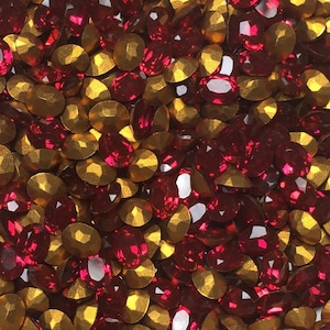 May include: A round container filled with a mix of red and gold-colored rhinestones. The rhinestones are oval-shaped and faceted, reflecting light. The container is white with a slightly textured surface. The rhinestones are likely used for crafts or embellishments.