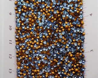 May include: Close-up of a collection of small, sparkling rhinestones. The rhinestones are a mix of light blue and gold colors. The gold rhinestones have a conical shape, while the blue ones are round with multiple facets. These are ideal for craft projects.