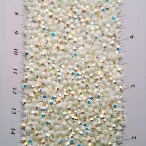 Könnte beinhalten: Nahaufnahme eines Haufens irisierender, weißer Strasssteine. Die kleinen, facettierten Strasssteine reflektieren verschiedene Farben. Ein Lineal mit Zentimetermarkierungen ist zur Größenbestimmung sichtbar.