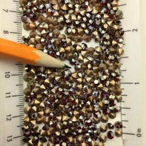 May include: A close-up image of a large quantity of small, round, iridescent rhinestones. The rhinestones are a mix of gold and red colors. A pencil is lying on top of the rhinestones, and a ruler is visible in the background.