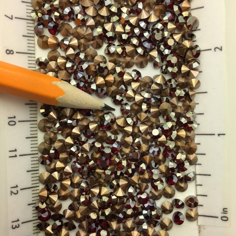 May include: A close-up image of a large quantity of small, round, iridescent rhinestones. The rhinestones are a mix of gold and red colors. A pencil is lying on top of the rhinestones, and a ruler is visible in the background.