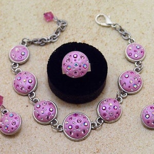 May include: A pink and silver jewelry set with a bracelet, earrings, and a ring. Each piece features a round, pink charm with a sparkly, iridescent design.
