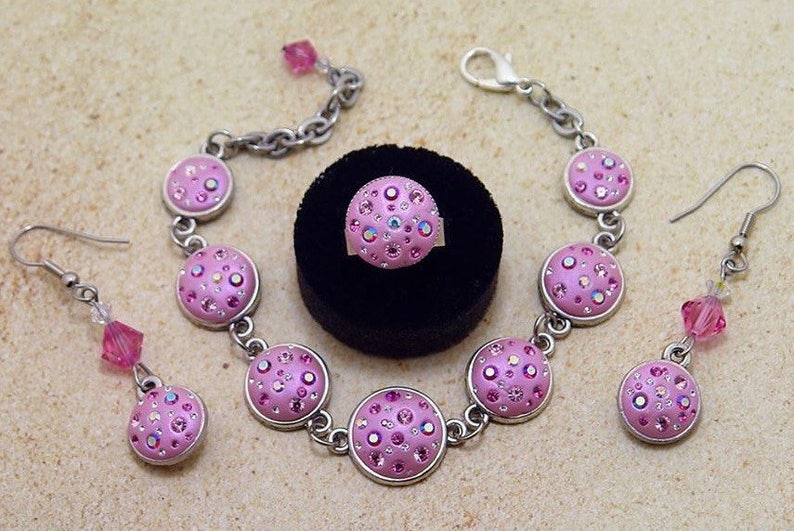 May include: A pink and silver jewelry set with a bracelet, earrings, and a ring. Each piece features a round, pink charm with a sparkly, iridescent design.