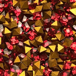 May include: A close-up image of a pile of red and gold pyramid-shaped rhinestones. The rhinestones are scattered randomly and are all the same size.