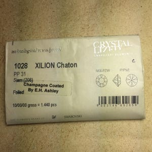 May include: A close-up of a white packaging label for Swarovski Xilion Chaton crystals. The label features the product number 1028, the color Siam (208), and the description "Champagne Coated Foiled by E.H. Ashley". The label also includes the text "10/00/00 gross = 1.440 pcs".
