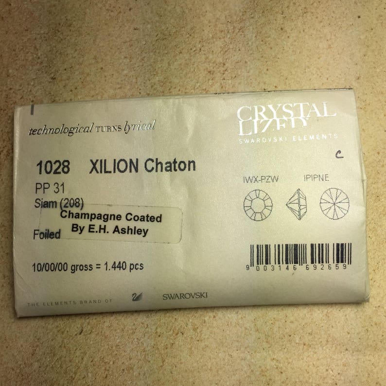 May include: A close-up of a white packaging label for Swarovski Xilion Chaton crystals. The label features the product number 1028, the color Siam (208), and the description "Champagne Coated Foiled by E.H. Ashley". The label also includes the text "10/00/00 gross = 1.440 pcs".
