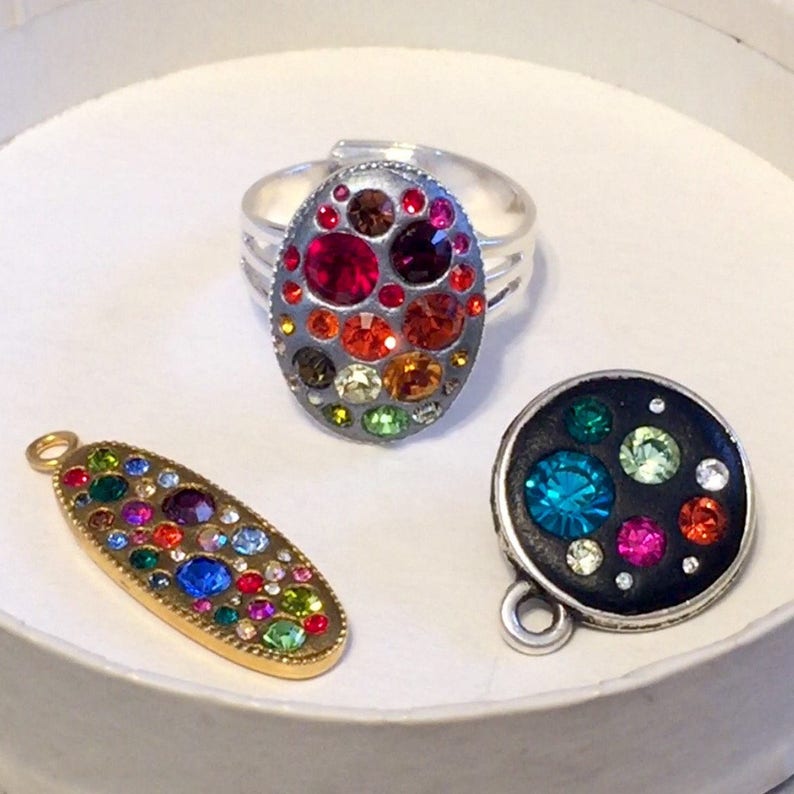 May include: Three oval-shaped jewelry pieces with a silver, gold, and black base. Each piece is adorned with colorful rhinestones of various sizes and colors.
