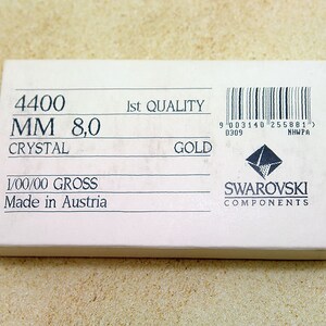 以下が含まれることがあります： 「4400 MM 8,0 CRYSTAL GOLD」と「Made in Austria」を含む黒い文字が書かれた白い箱。箱にはバーコードとスワロフスキーコンポーネンツのロゴも表示されています。