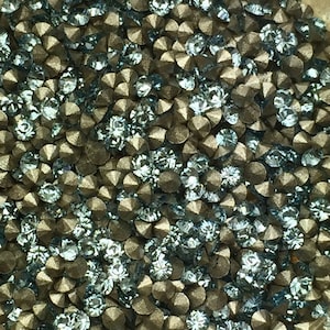 May include: A close-up image of a large quantity of small, round, light blue rhinestones with a silver metallic finish. The rhinestones are scattered randomly and appear to be loose.