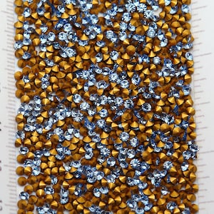 May include: Close-up of a collection of small, faceted rhinestones. The rhinestones are a mix of light blue and gold colours, creating a sparkling effect. The gems are scattered across a white surface, suggesting a craft or jewellery-making project.