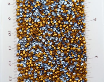May include: Close-up of a collection of small, faceted rhinestones. The rhinestones are a mix of light blue and gold colors, creating a sparkling effect. The gems are scattered across a white surface, suggesting a craft or jewelry-making project.