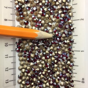 May include: A close-up image of a large quantity of small, round, red and gold rhinestones. The rhinestones are scattered on a white surface with a ruler in the background. The ruler is marked in inches.