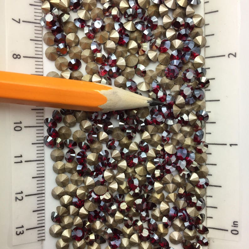 May include: A close-up image of a large quantity of small, round, red and gold rhinestones. The rhinestones are scattered on a white surface with a ruler in the background. The ruler is marked in inches.
