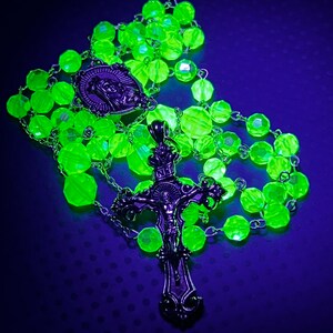Puede incluir: Un rosario verde ne&oacute;n con cuentas facetadas y detalles met&aacute;licos plateados. El rosario presenta un colgante de crucifijo con una representaci&oacute;n de Jes&uacute;s y un medall&oacute;n con una figura religiosa. Las cuentas est&aacute;n ensartadas en una cadena plateada.