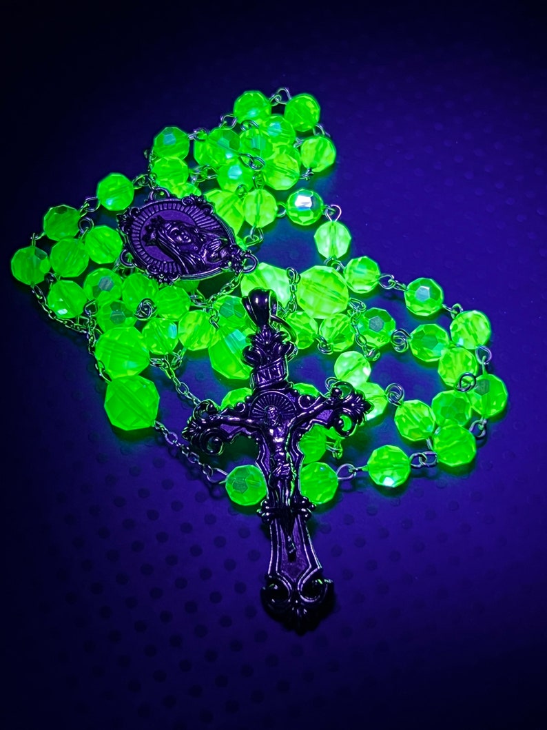 Puede incluir: Un rosario verde ne&oacute;n con cuentas facetadas y detalles met&aacute;licos plateados. El rosario presenta un colgante de crucifijo con una representaci&oacute;n de Jes&uacute;s y un medall&oacute;n con una figura religiosa. Las cuentas est&aacute;n ensartadas en una cadena plateada.